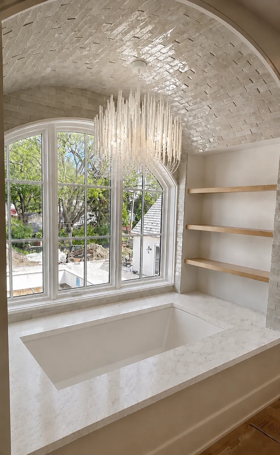 A tub under an arched window, with a glazed tile vault, a crystal chandelier and open shelves.